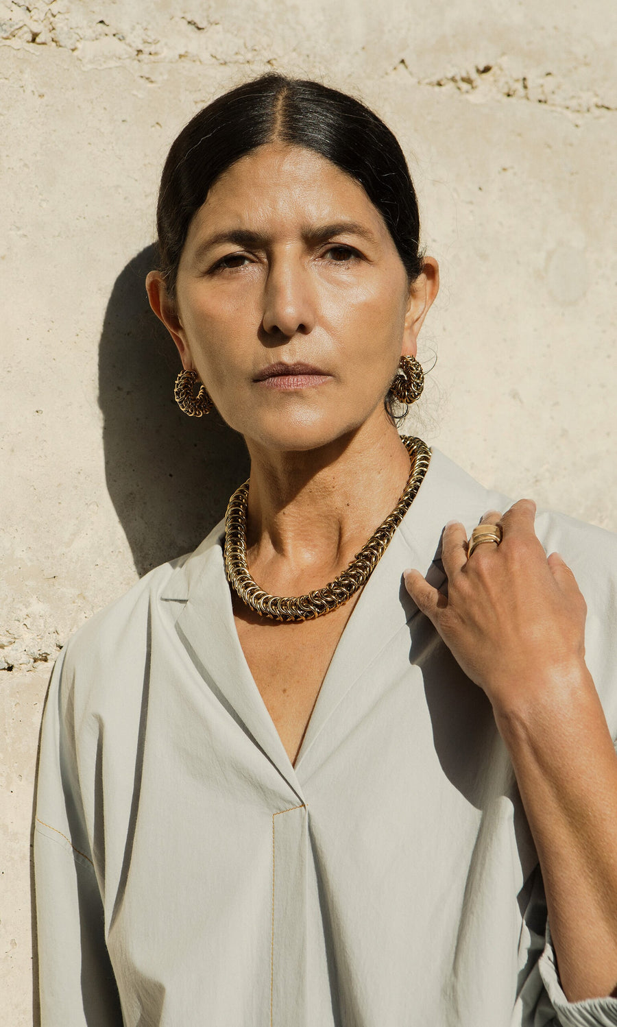 Plümo- Woman with dark hair in a bun, wearing Carew Hoops and a light blouse, stands against a beige wall.