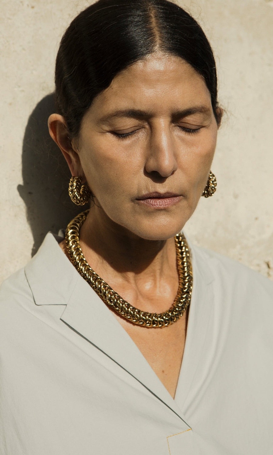 Plümo- Woman with closed eyes wears the Kibbin Necklace, gold-plated, and earrings in a light blouse against a sunlit wall.