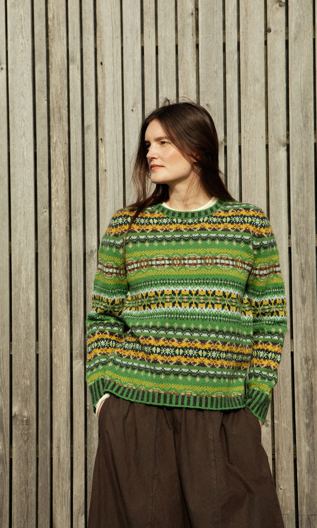 Plümo- Woman in a green St Kilda Jumper and brown skirt stands before a wooden plank wall, looking to the side.