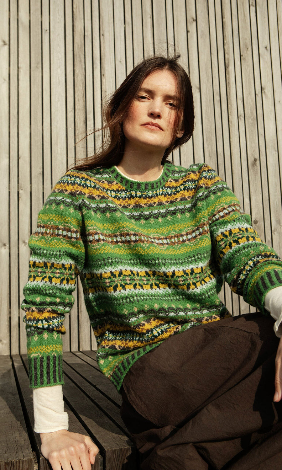 Plümo- Woman in a St Kilda Jumper sits outdoors by a wooden wall, looking towards the camera in sunlight.