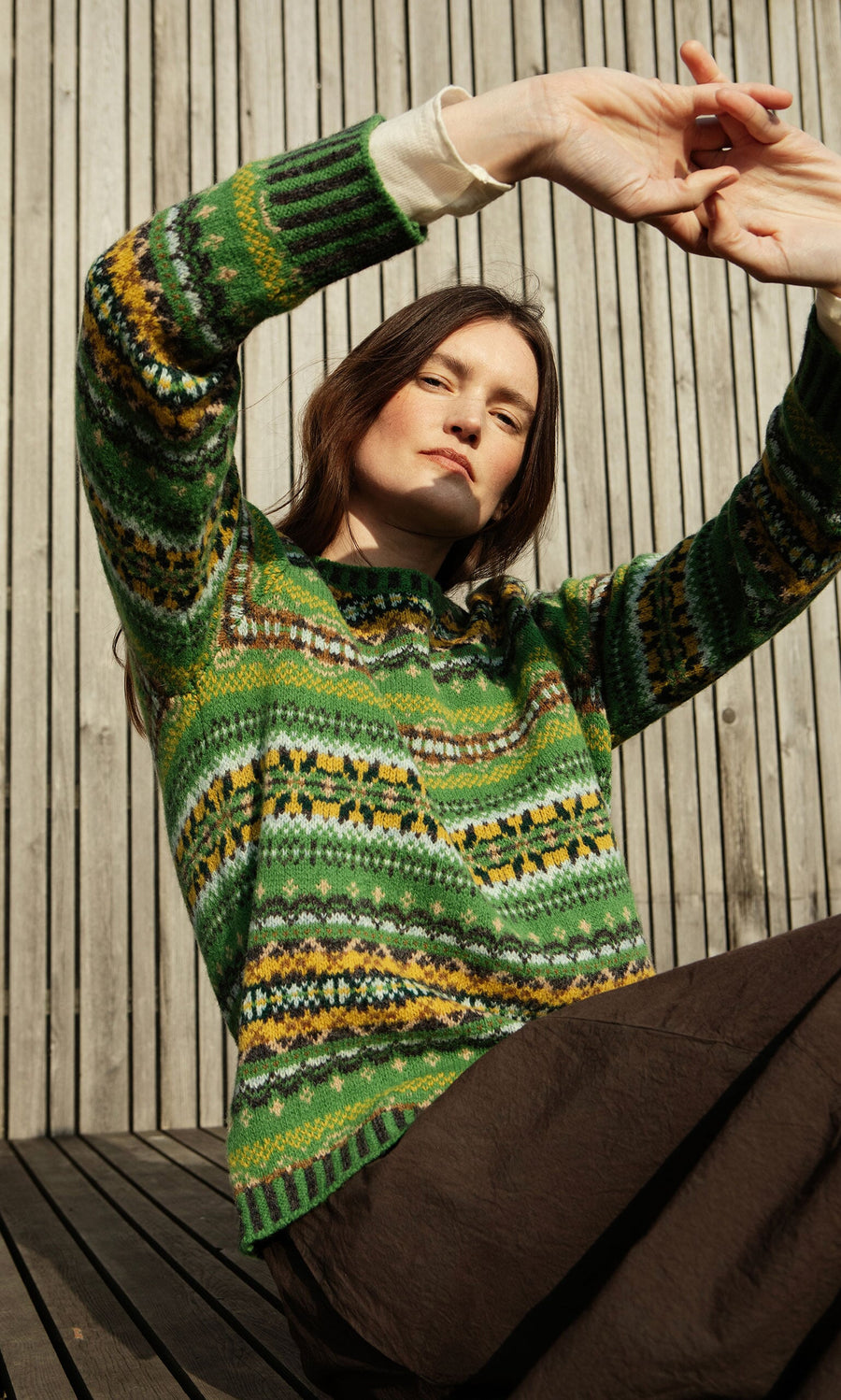Plümo- Woman in a St Kilda Jumper sits on wooden decking, arms raised, with a wooden wall as the backdrop.