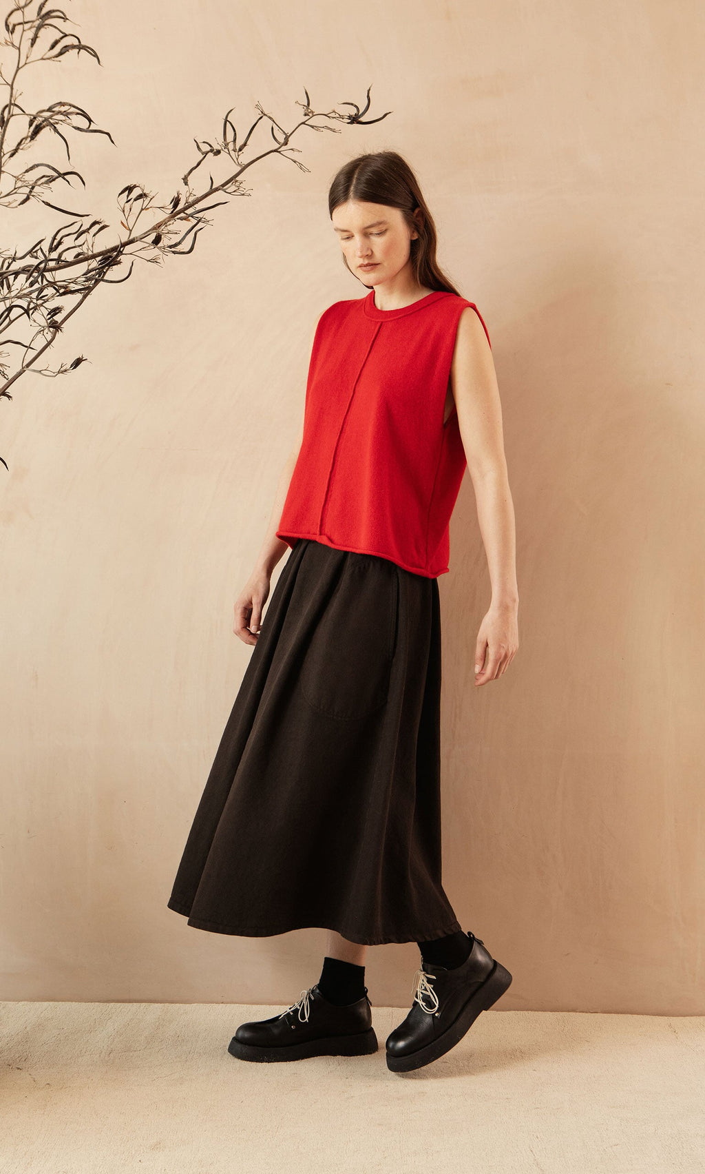 Plümo- A woman in a red sleeveless top and the Allard Skirt stands beside a branch against a beige background.