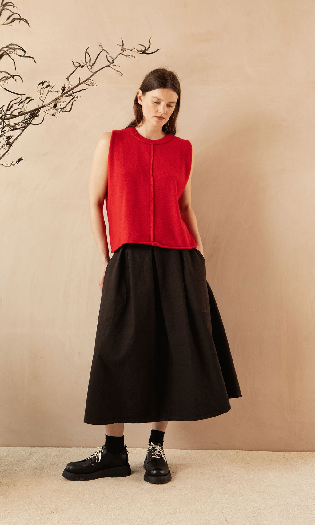 Plümo- Woman in a sleeveless red top and the Allard Skirt stands against a beige wall, looking down with hands in pockets.