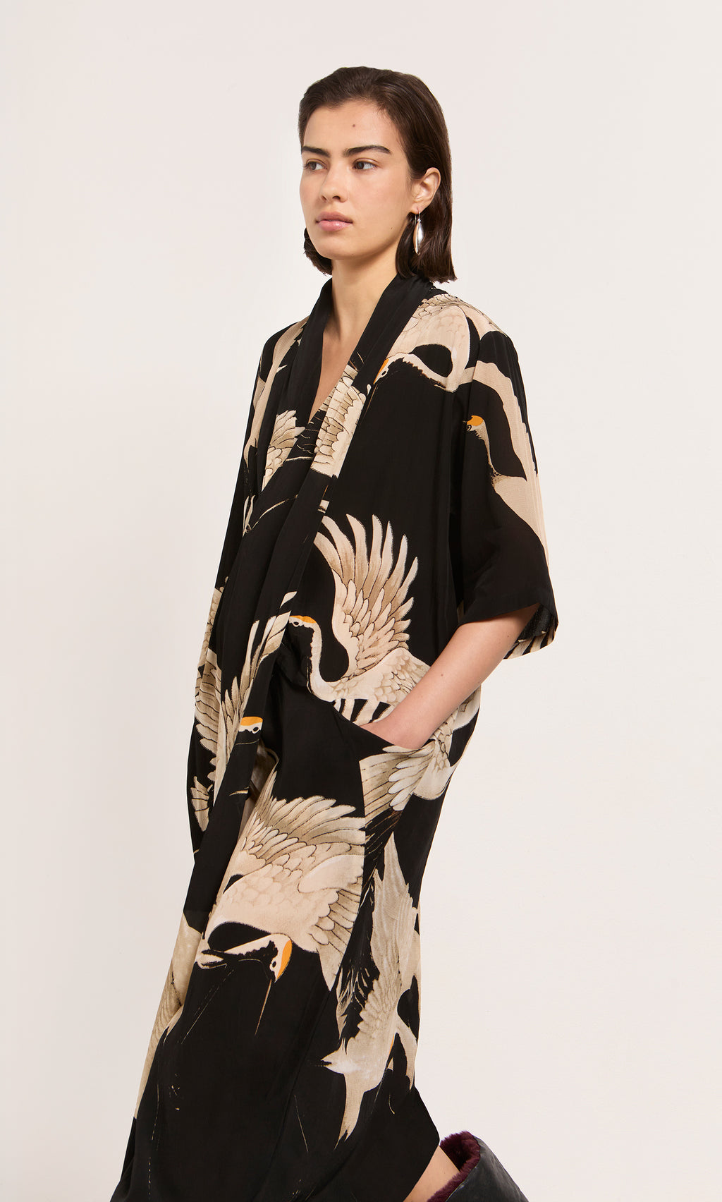 Plümo  Woman in the Stork Twist Dress with crane prints, hands in pockets, stands against a plain light background.