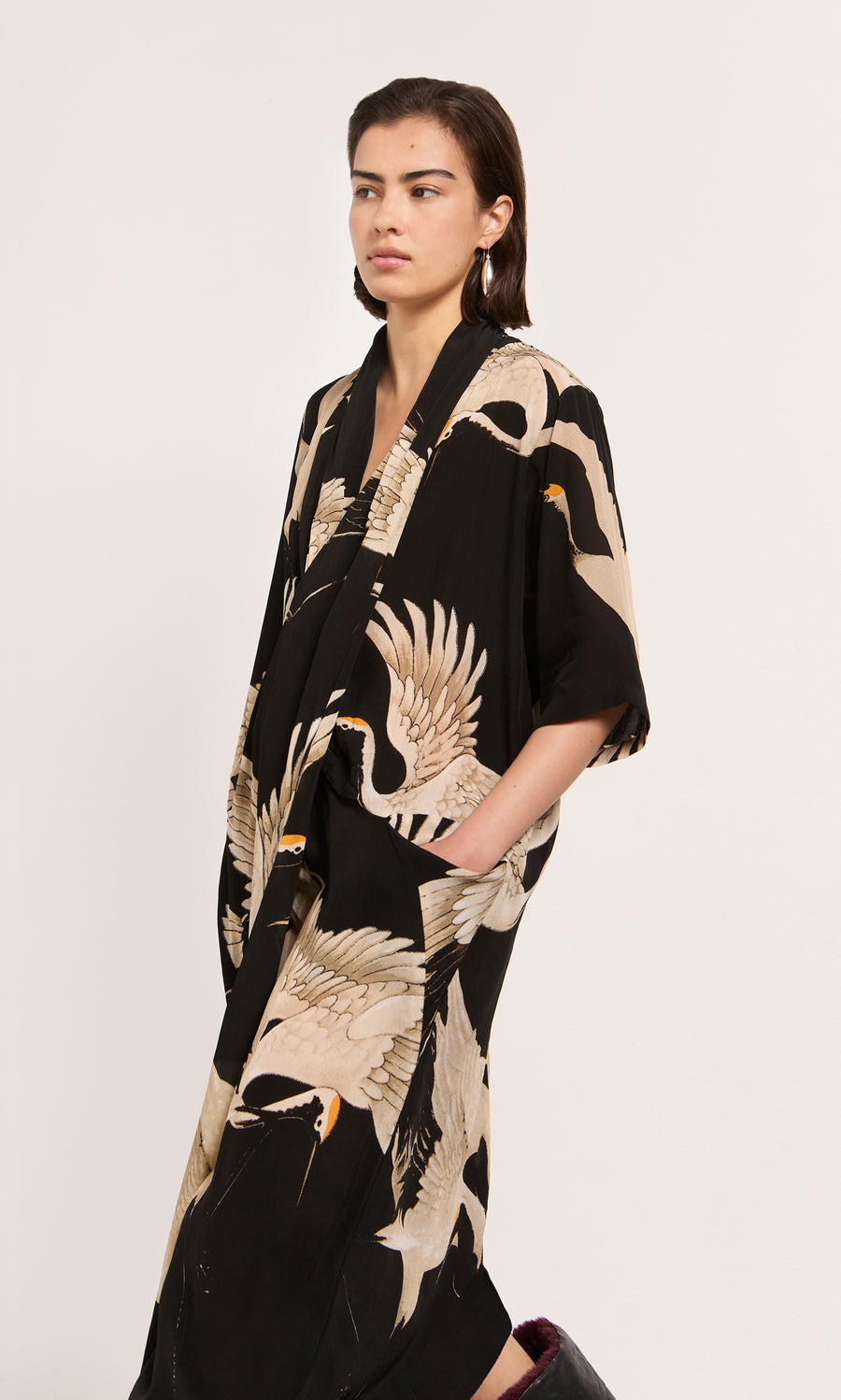Plümo  Woman in the Stork Twist Dress with crane prints, hands in pockets, stands against a plain light background.