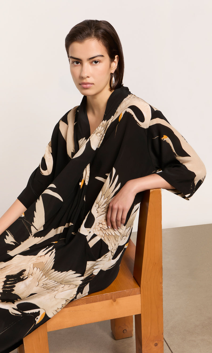 Plümo  Woman in the Stork Twist Dress sits on a wooden chair, facing the camera in vintage-inspired style.