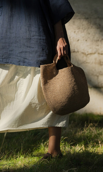 Plümo- A person in a navy top and white skirt stands on grass, holding the Tara Straw Bag in sunlight.