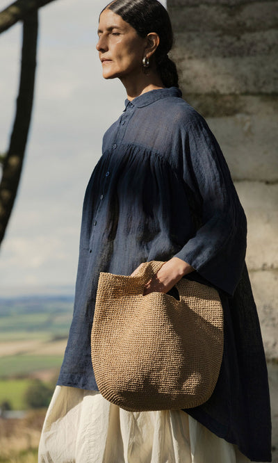 Plümo- A woman holds the Tara Straw Bag while wearing a loose navy top and white skirt near a stone wall outdoors.