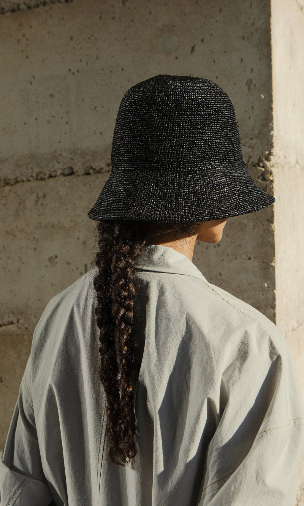 Plümo- A person with a long braid wears a Straw Bucket Hat and light shirt, standing with their back to a concrete wall.