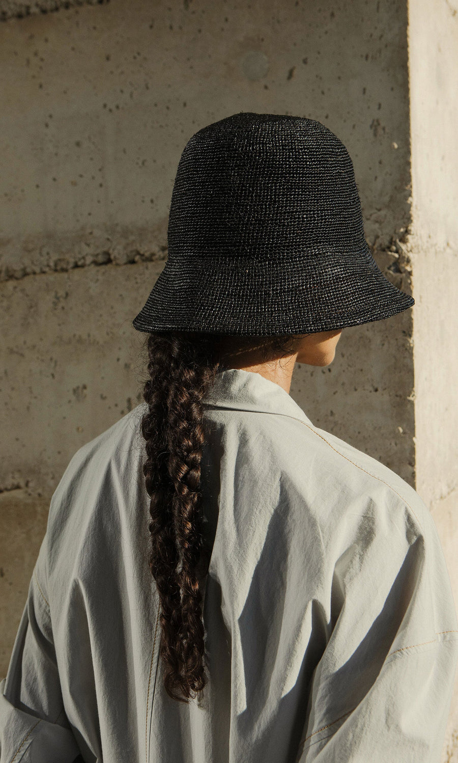 Plümo- A person with a long braid wears a Straw Bucket Hat and light shirt, standing with their back to a concrete wall.