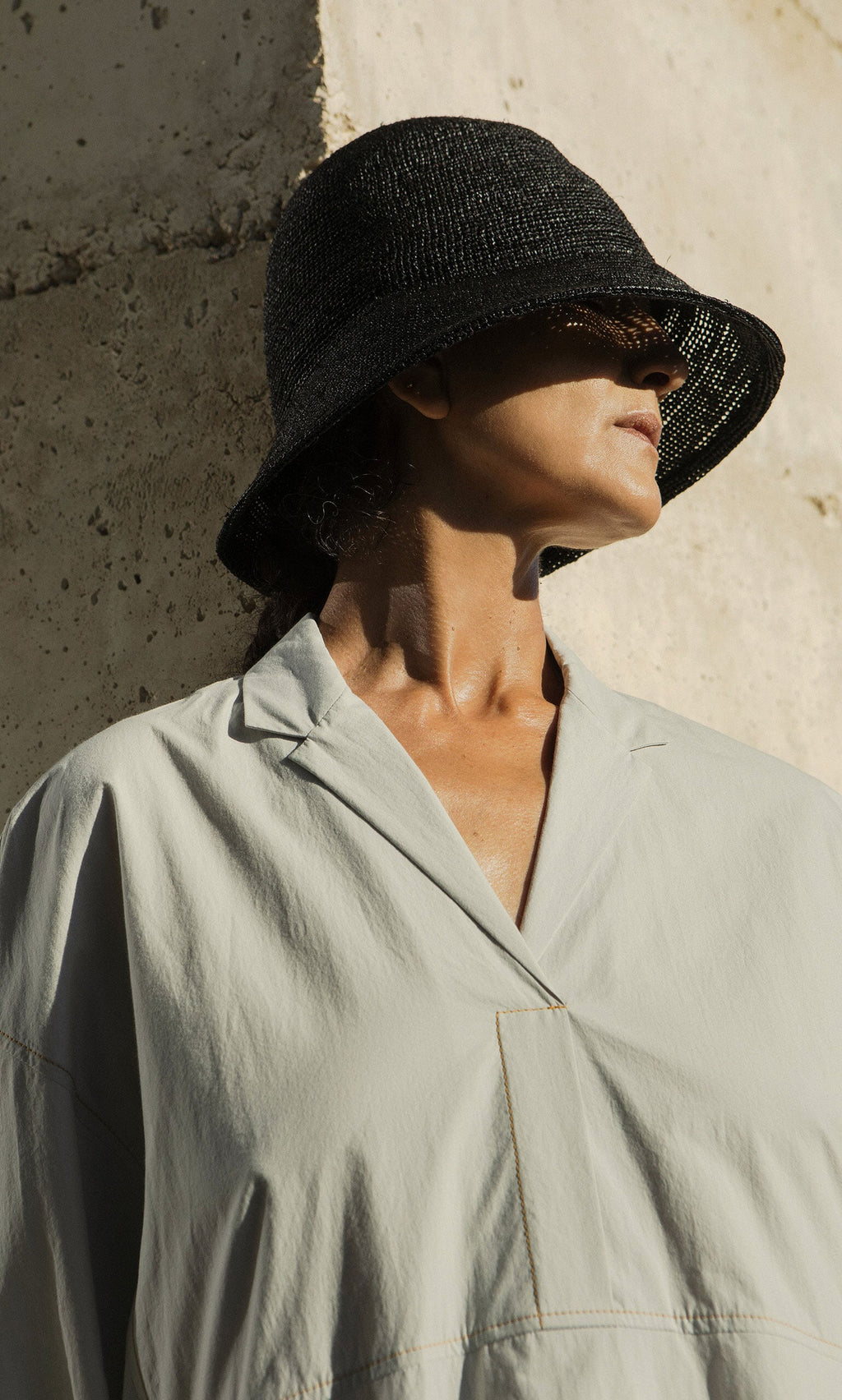 Plümo- Person in a light gray shirt wears a Straw Bucket Hat, standing against a textured beige wall with face partly in shadow.