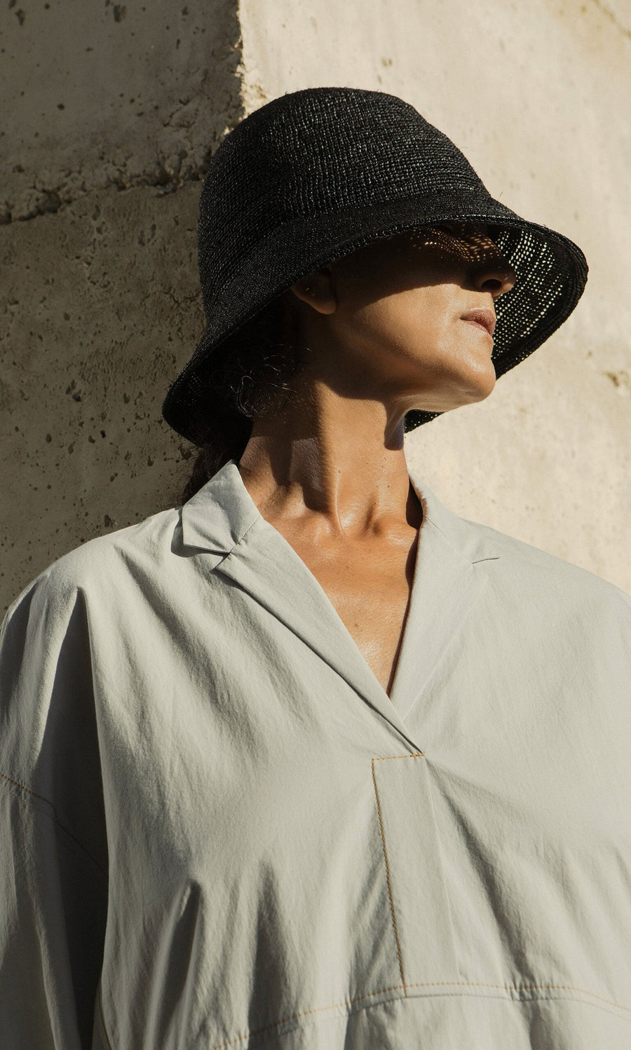 Plümo- Person in a light gray shirt wears a Straw Bucket Hat, standing against a textured beige wall with face partly in shadow.