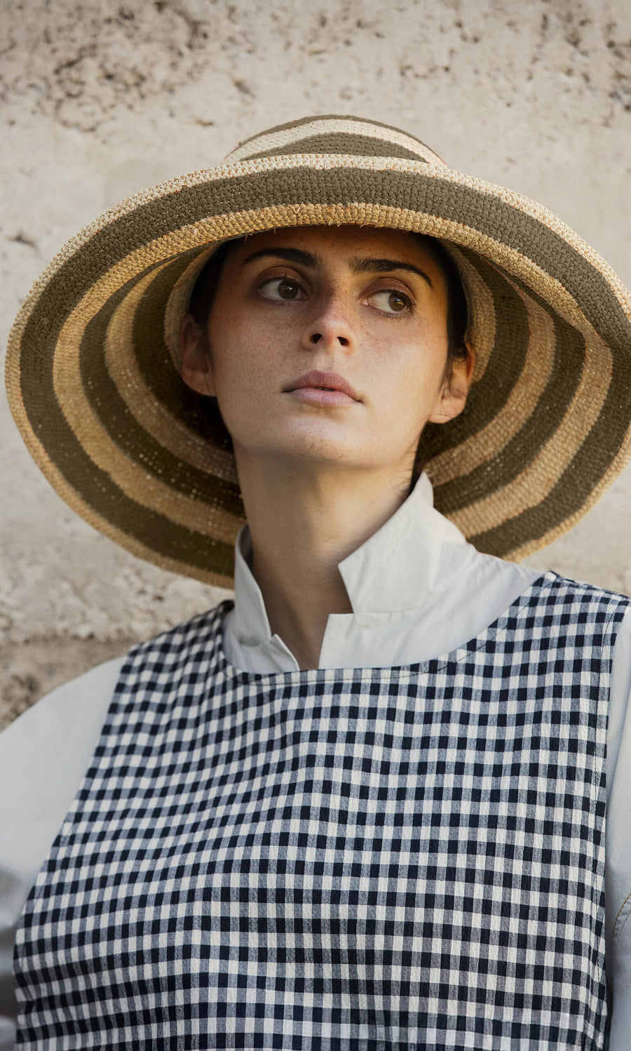 Plümo- Woman in a Stripe Sun Hat and checkered dress looks away, standing against a textured stone wall.