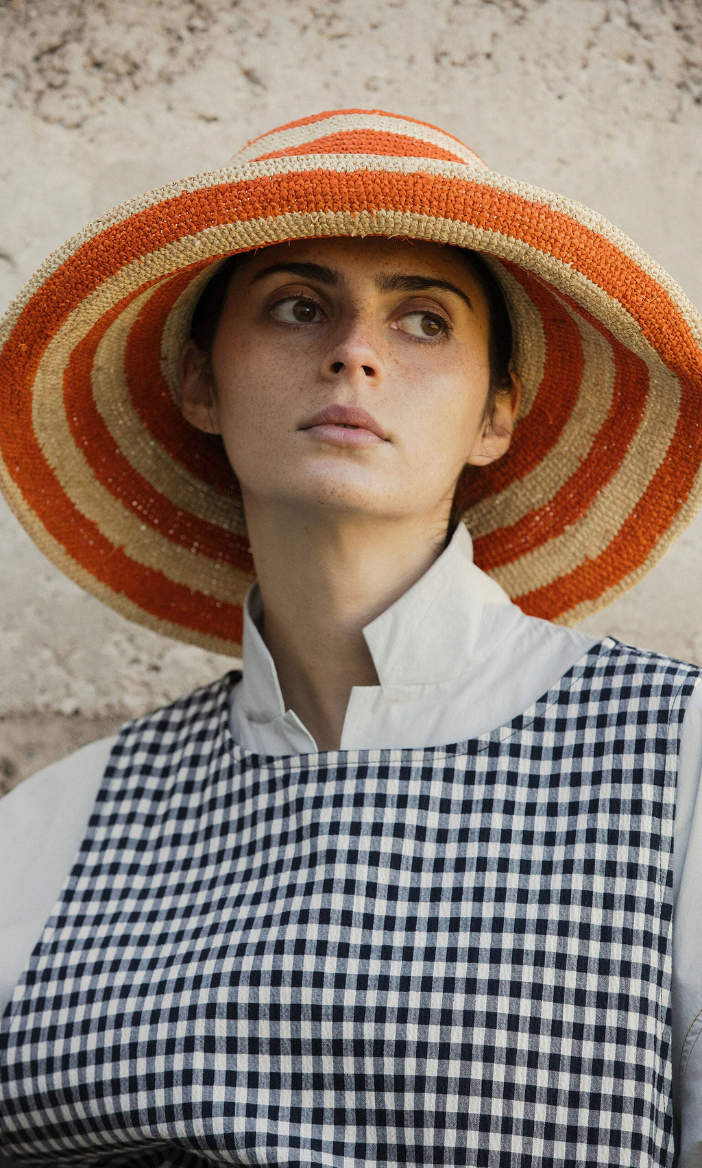Plümo- Wearing the Orange Stripe Sunhat, a person looks to the side against a textured wall.