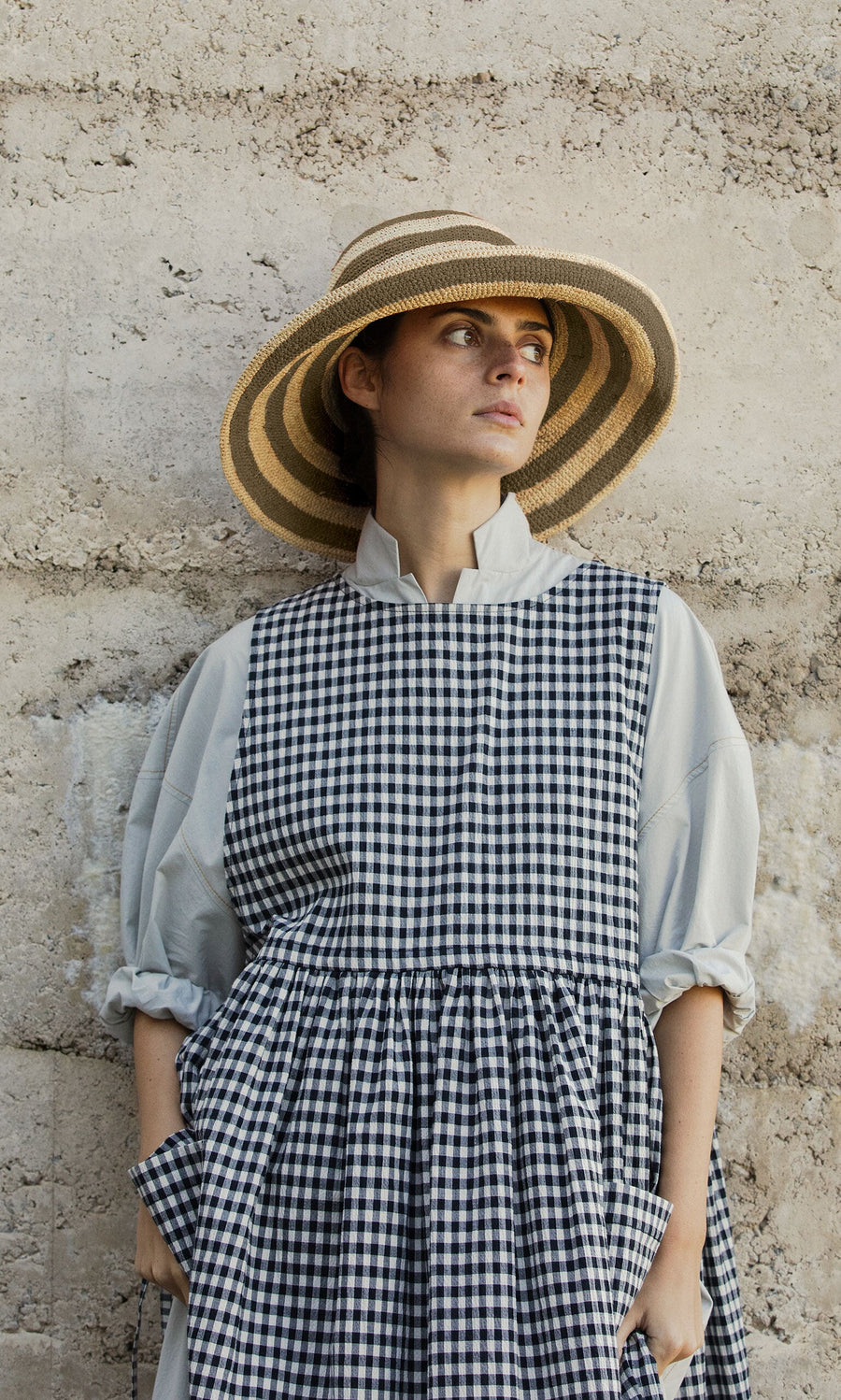 Plümo- Woman in a Stripe Sun Hat and checkered dress stands by a stone wall, looking to the side.