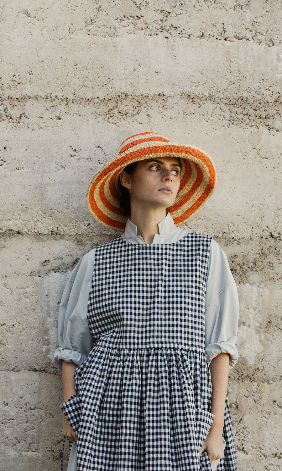 Plümo- Woman in a checkered dress and Orange Stripe Sunhat stands by a textured stone wall, gazing to the side.