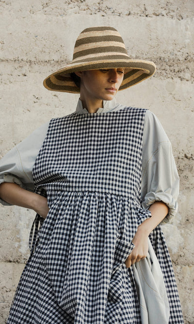 Plümo- Woman wearing a Stripe Sun Hat and a black-and-white checkered dress stands against a textured stone wall.