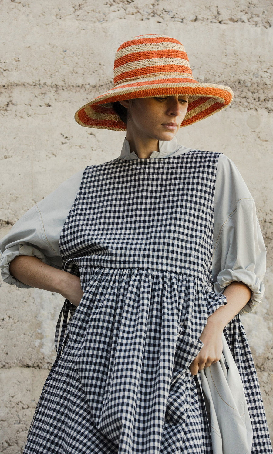 Plümo- Woman wearing the Orange Stripe Sunhat and a checkered dress, standing against a stone wall.