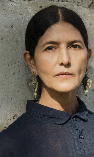 Plümo- Woman with dark hair wears Sunfish Earrings and a dark shirt, standing by a textured concrete wall.