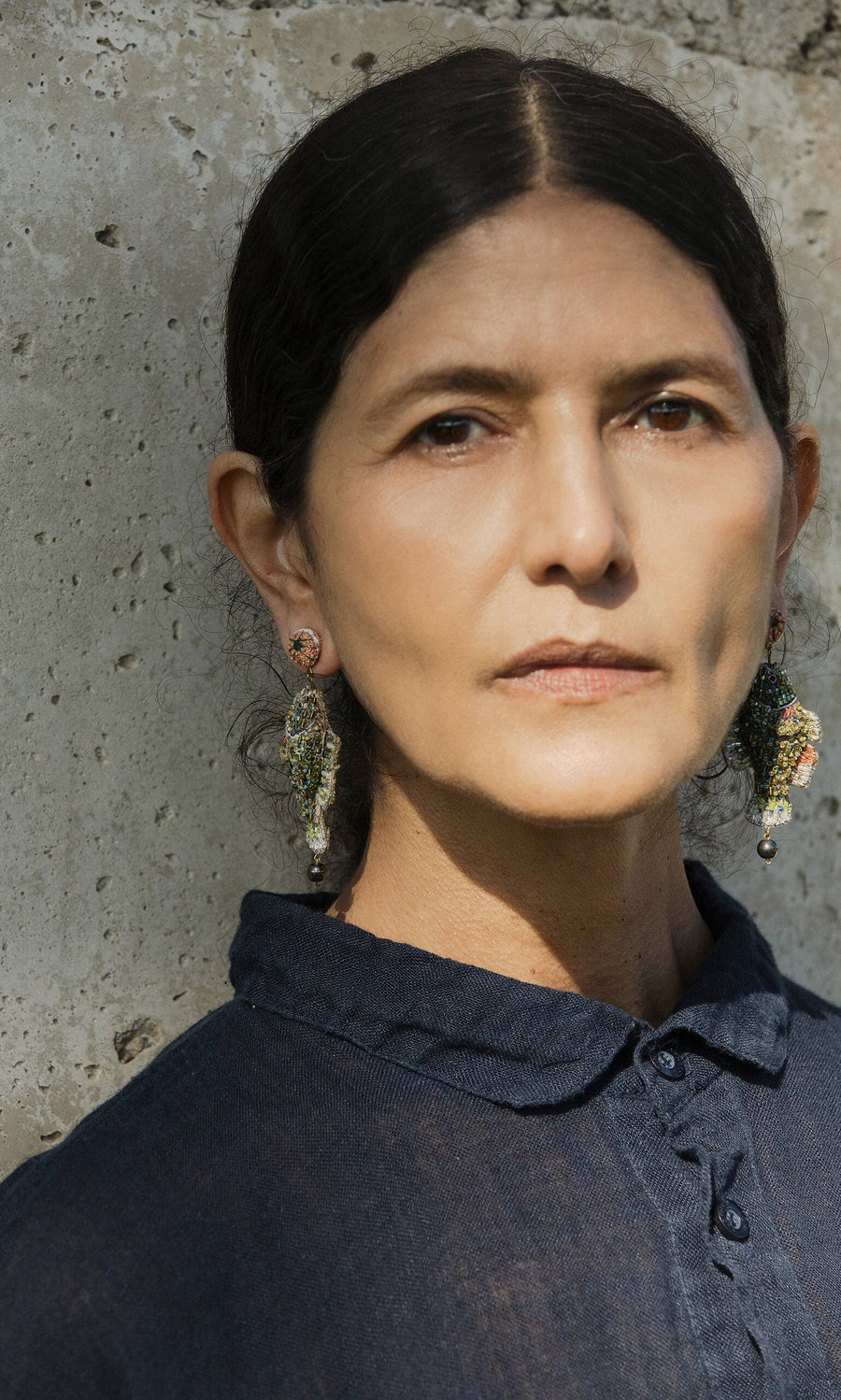 Plümo- Woman with dark hair wears Sunfish Earrings and a dark shirt, standing by a textured concrete wall.