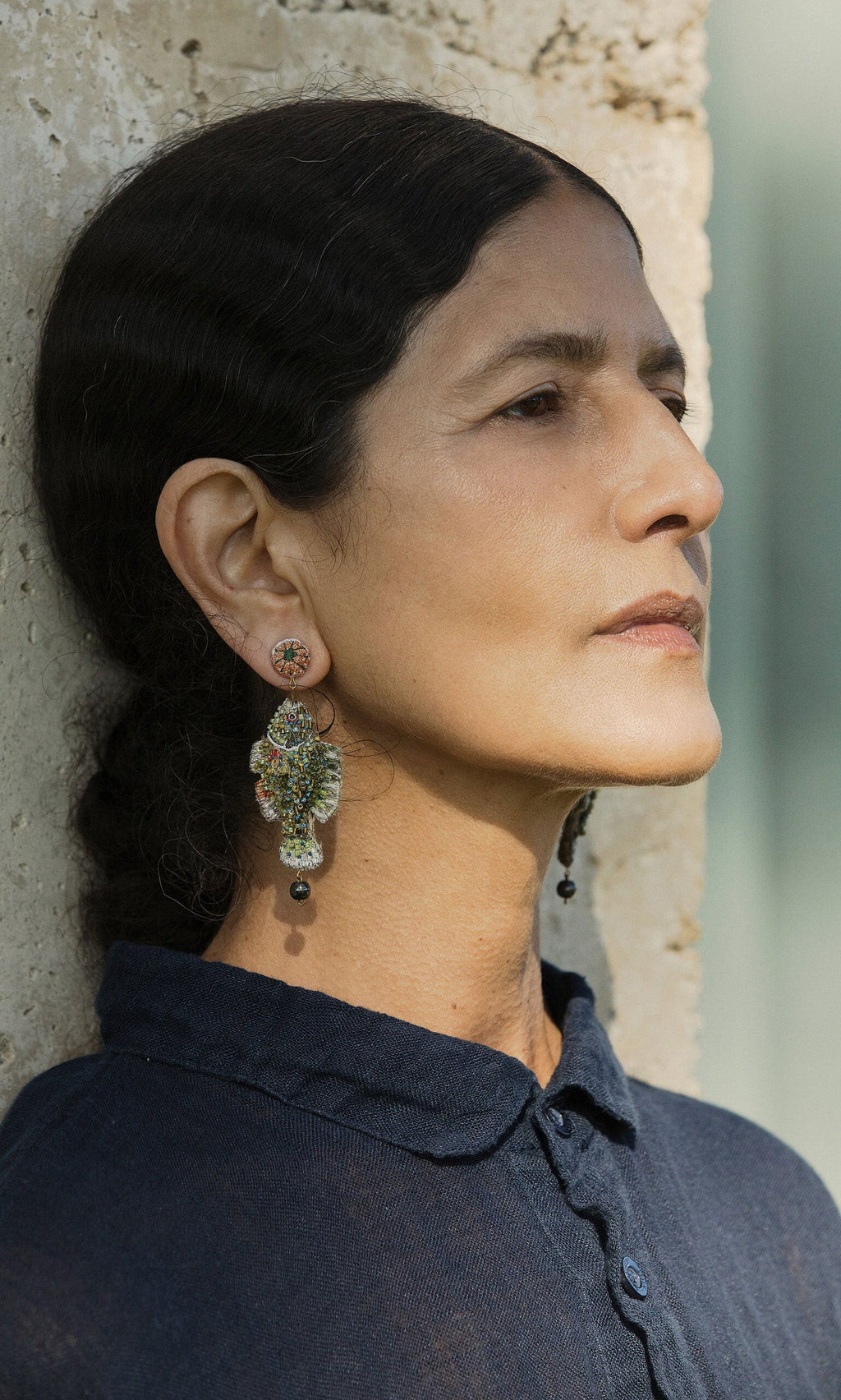 Plümo- Woman in profile, wearing a navy shirt and Sunfish Earrings, stands by a textured stone wall.