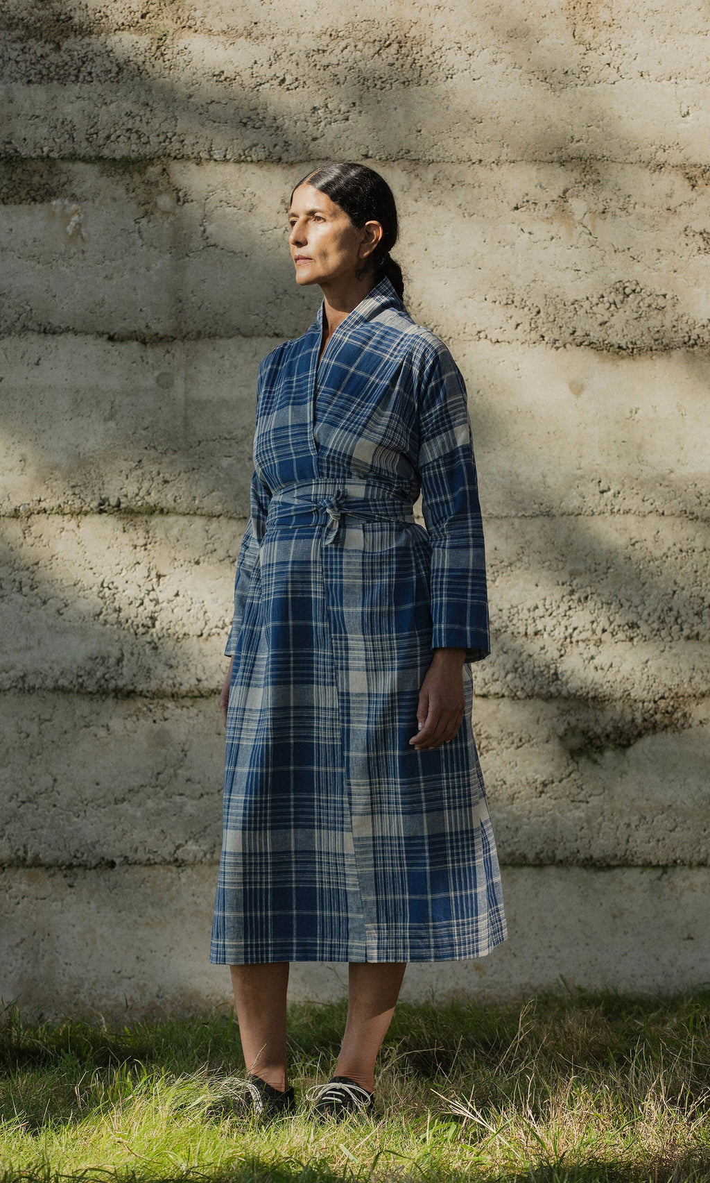 Plümo- Woman in the Wrap Tie - Check dress made from Japanese Banshu-ori stands on grass, looking to the side.