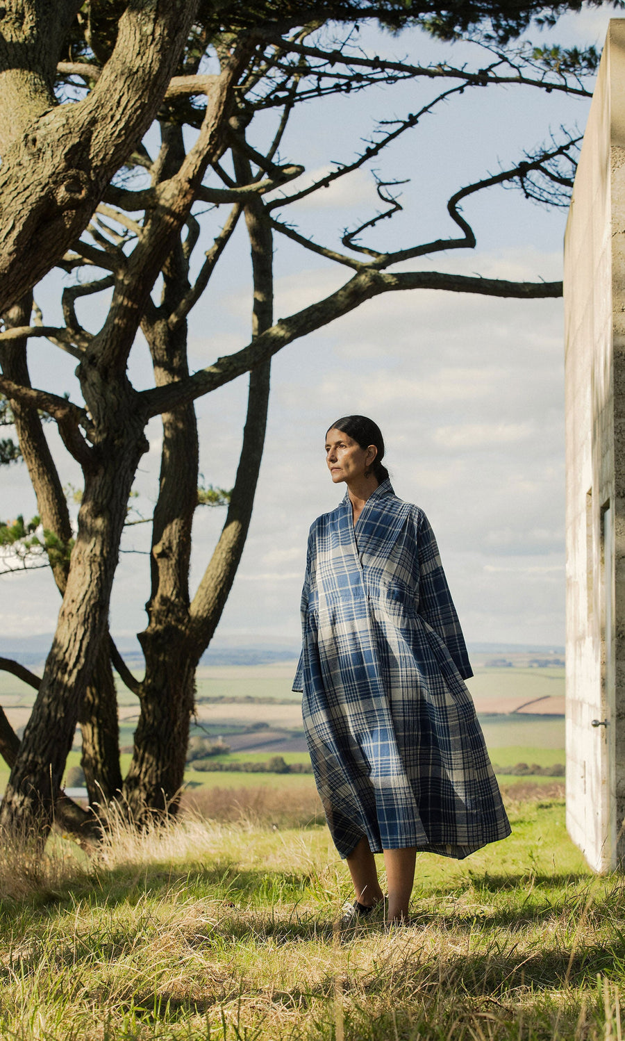 Plümo- A person wears the Tarkhan Dress - Check, standing outdoors by a tree with open fields and sky behind them.