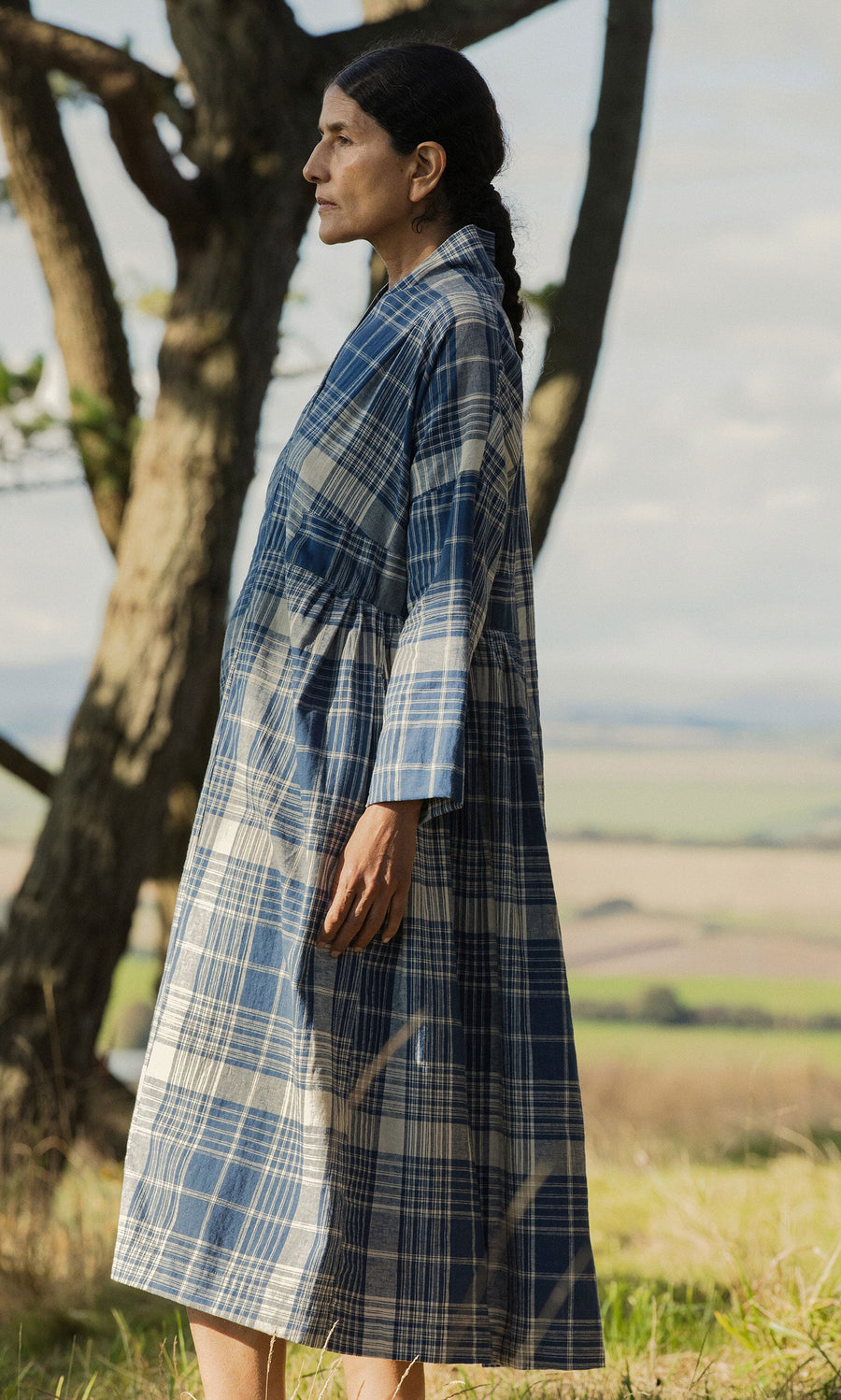 Plümo- Woman in a Tarkhan Dress - Check stands outdoors in profile, with trees and fields in the background.