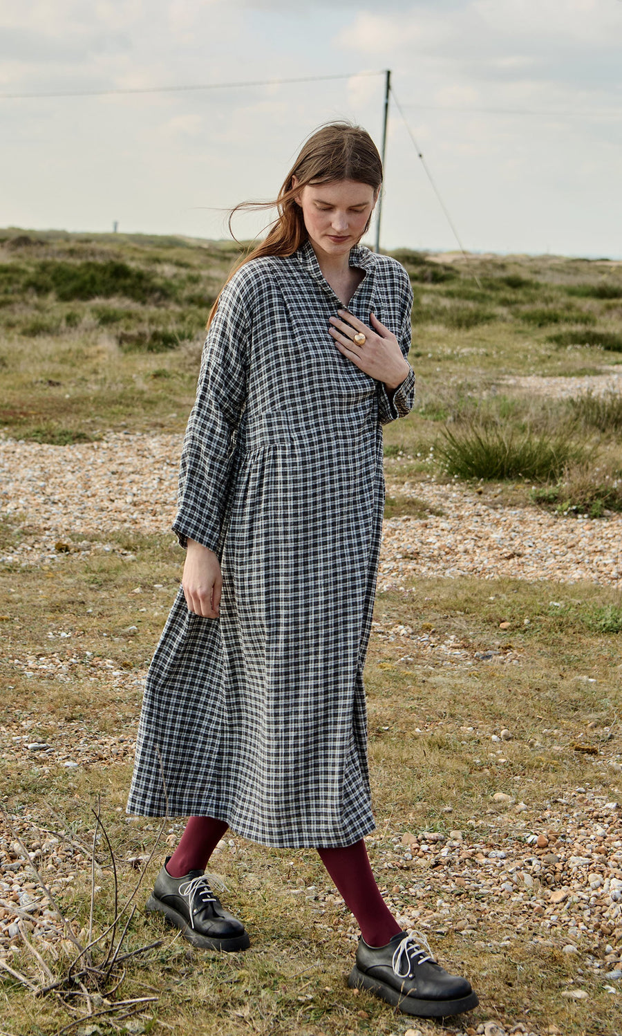 Plümo- A woman in the Tarkhan Dress - Plaid walks on a grassy field, paired with black shoes and burgundy tights.