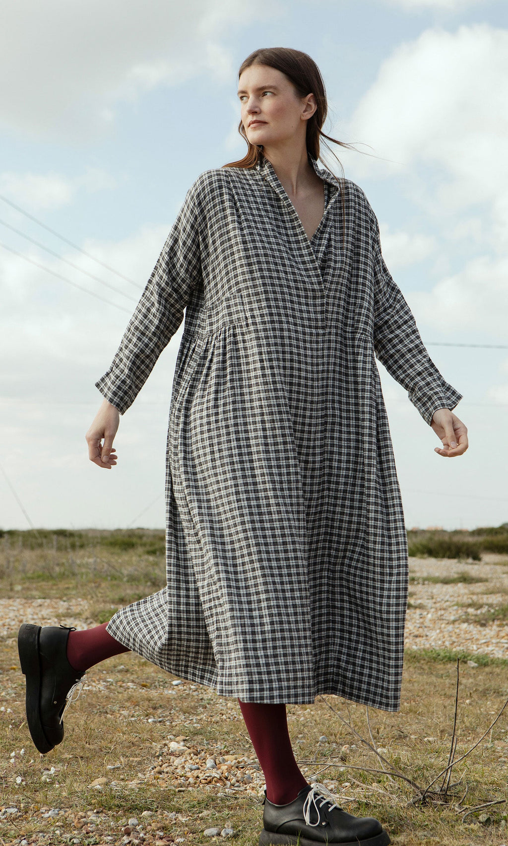 Plümo- A woman in the Tarkhan Dress - Plaid with black shoes walks outdoors on rocky ground in North London.