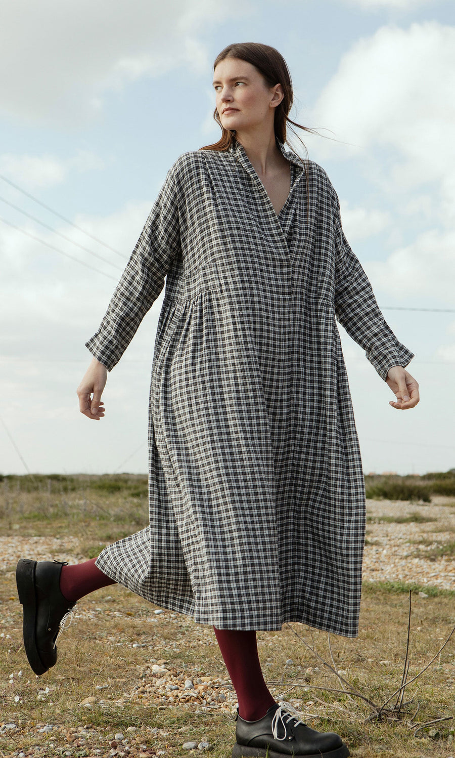Plümo- A woman in the Tarkhan Dress - Plaid with black shoes walks outdoors on rocky ground in North London.
