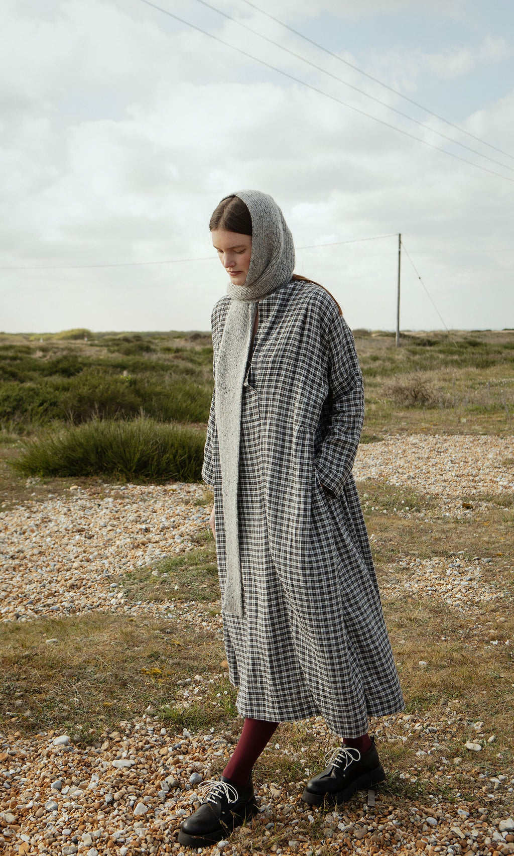 Plümo- Woman in the Tarkhan Dress - Plaid with a wrap-over V neck stands outdoors on a gravel path in North London.