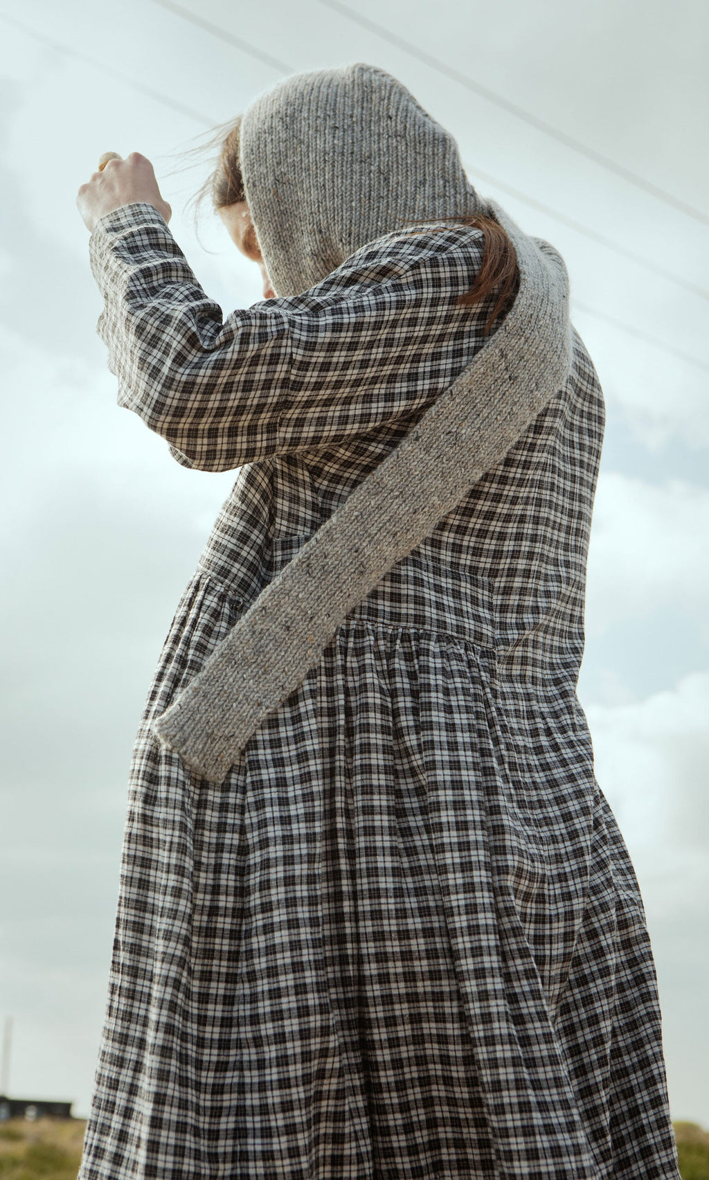 Plümo- A person in the Tarkhan Dress - Plaid with a knitted hooded scarf stands outdoors in North London, arm raised to the sky.