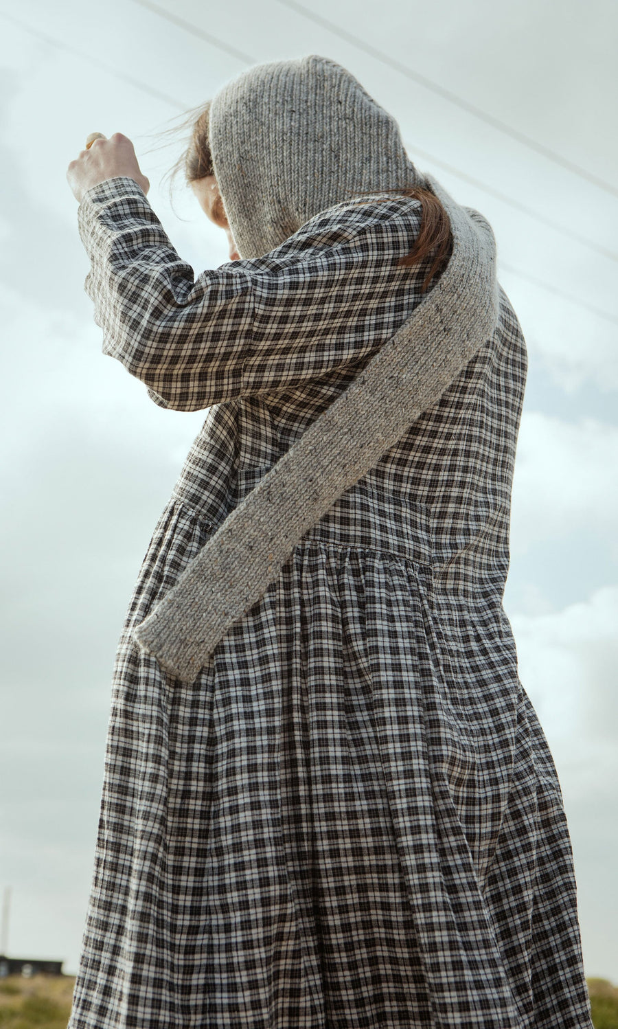 Plümo- A person in the Tarkhan Dress - Plaid with a knitted hooded scarf stands outdoors in North London, arm raised to the sky.