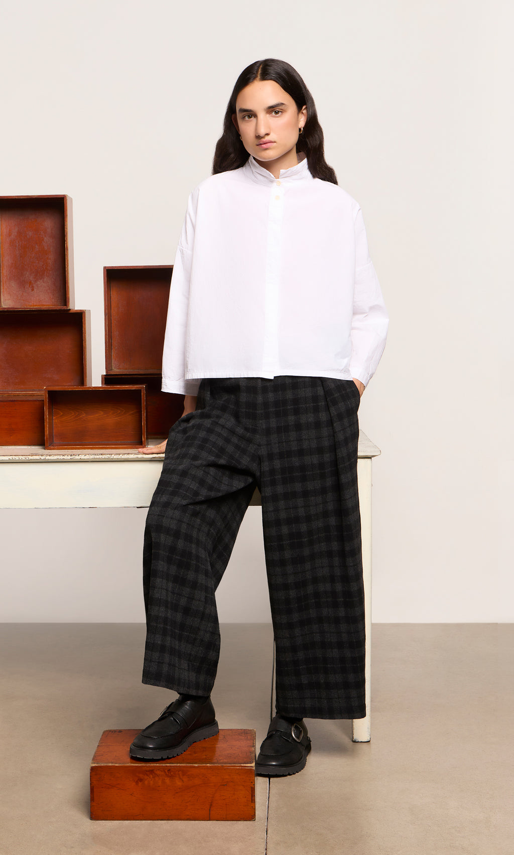 Plümo  Person in a white shirt and Tartan Flannel Pants stands by boxes and a table, looking at the camera.