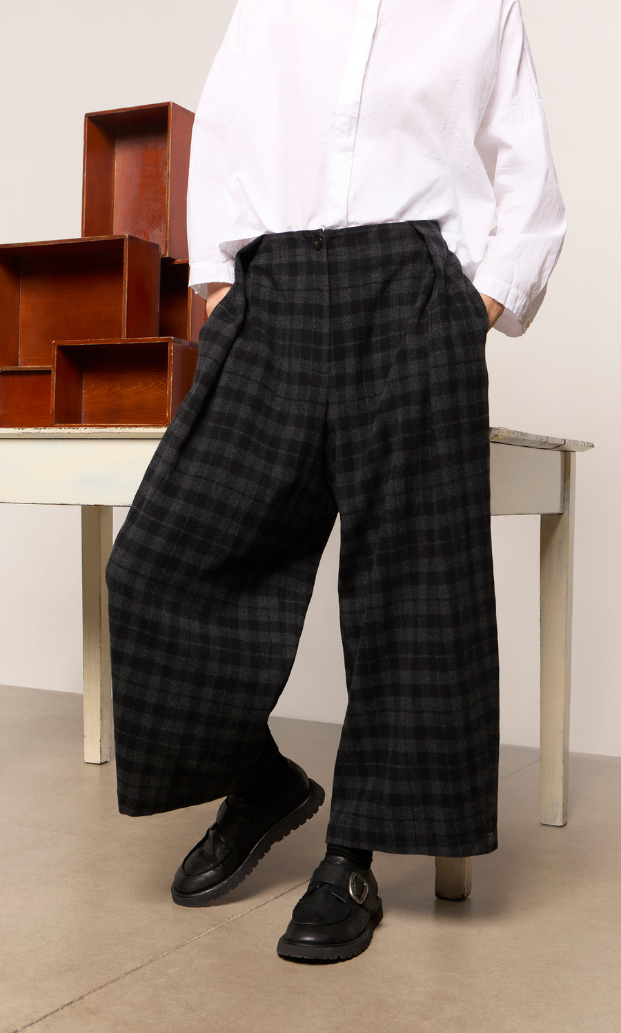 Plümo  Person in Tartan Flannel Pants, white shirt, and black shoes stands by a table and stacked boxes.