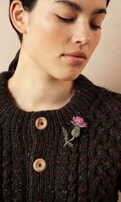 Plümo- Woman wears a dark knit sweater with large buttons and a Thistle Brooch pinned to the chest.