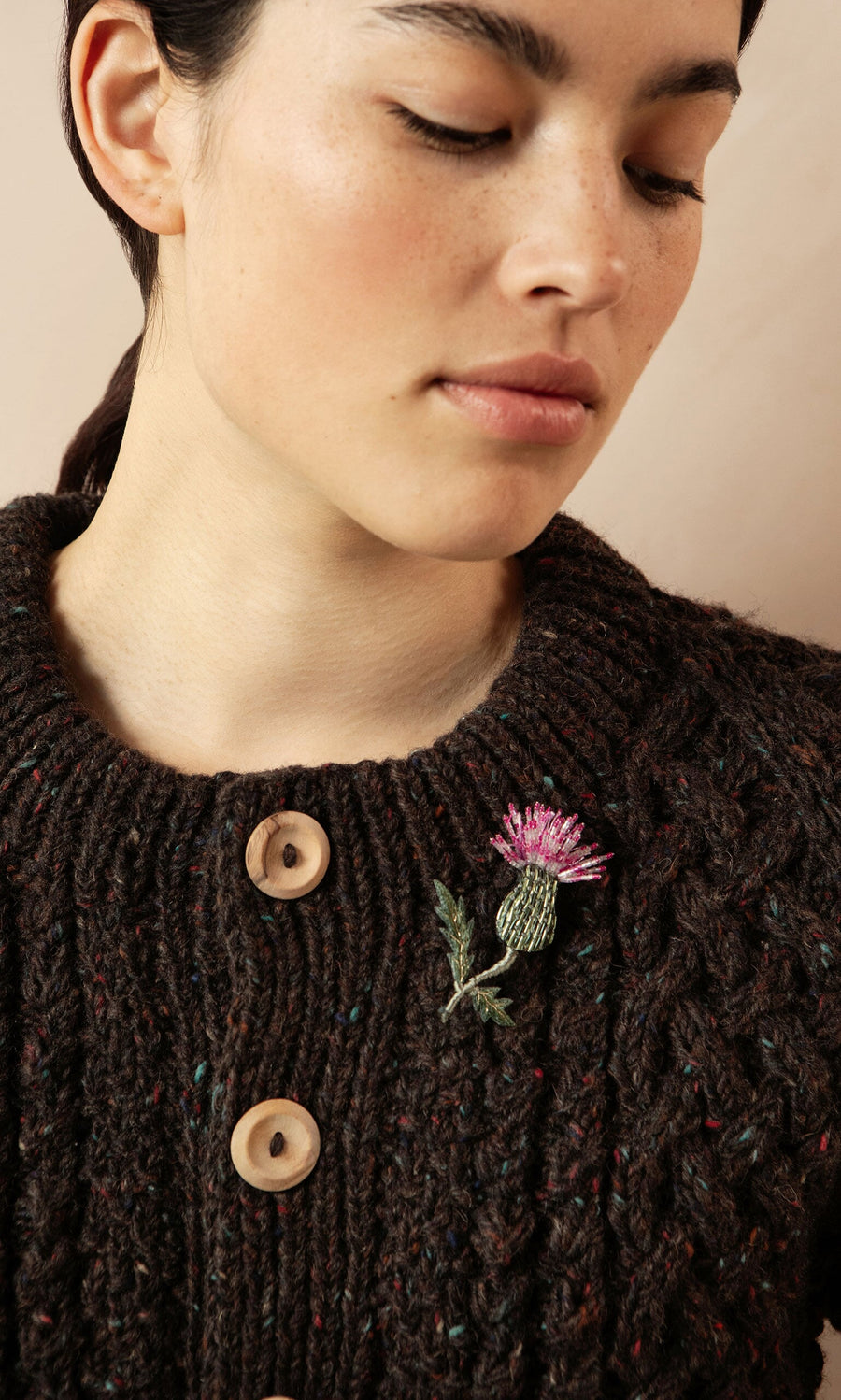 Plümo- Woman wears a dark knit sweater with large buttons and a Thistle Brooch pinned to the chest.