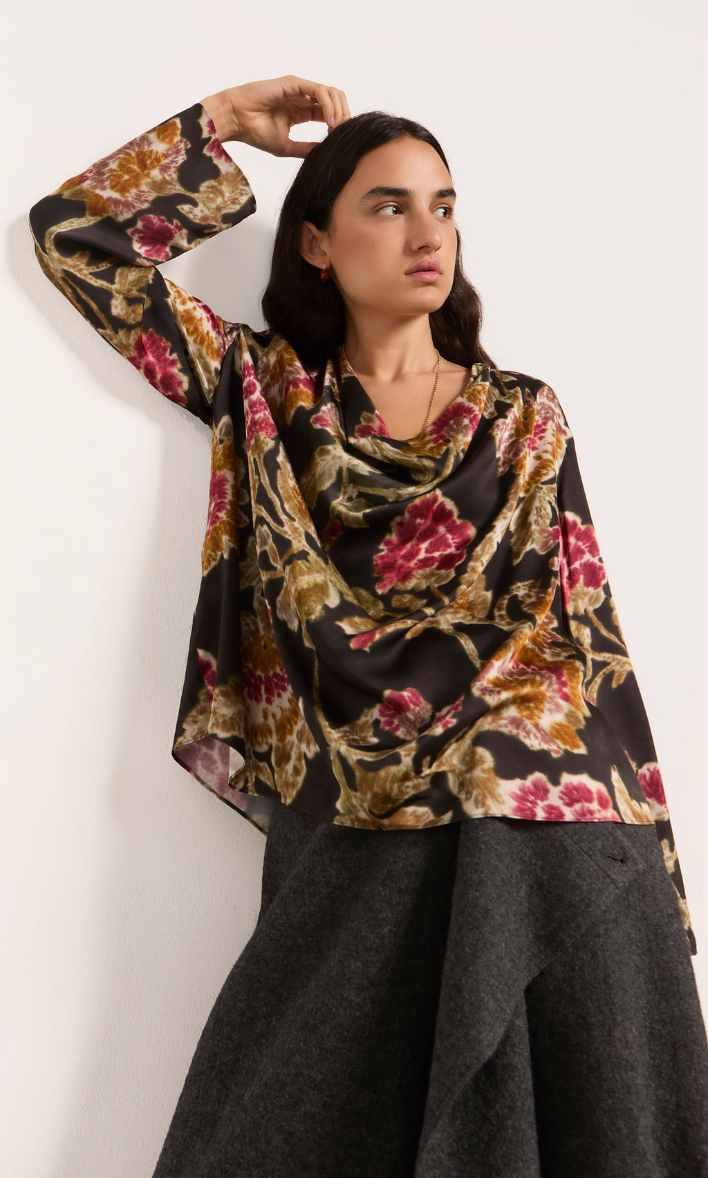 Plümo  Woman in a Tokley Blouse with a blurry floral print leans against a white wall, gazing slightly to the side.