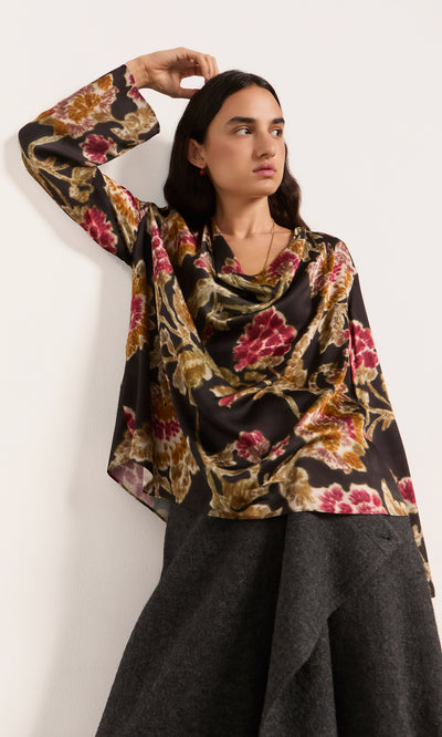 Plümo  Woman in a Tokley Blouse with a blurry floral print leans against a white wall, gazing slightly to the side.