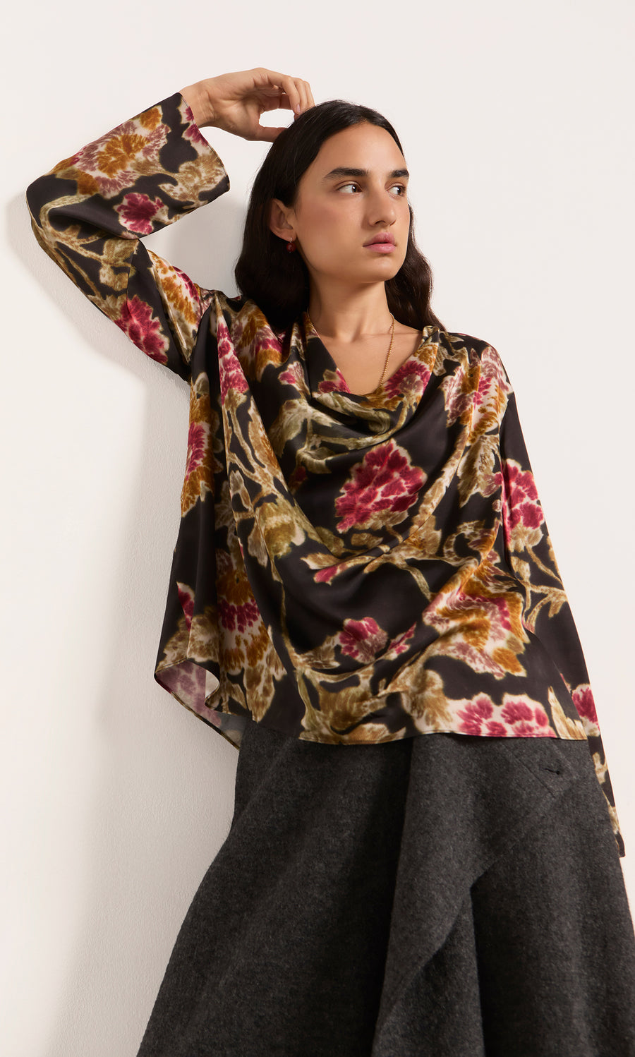 Plümo  Woman in a Tokley Blouse with a blurry floral print leans against a white wall, gazing slightly to the side.