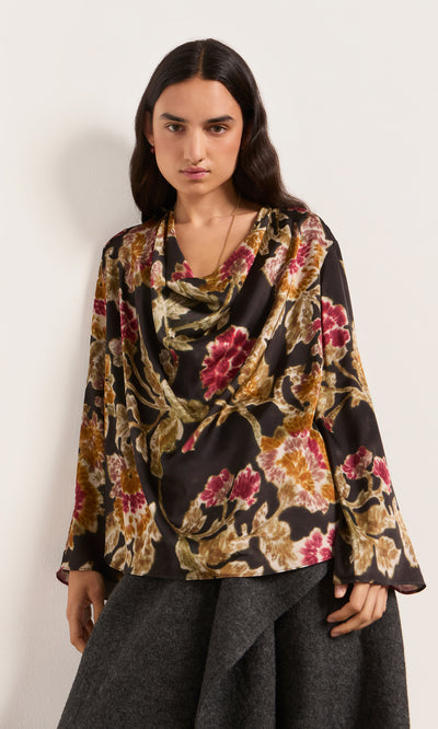 Plümo  Woman in a Tokley Blouse with blurry floral print, long sleeves, and gray skirt, standing against a plain backdrop.