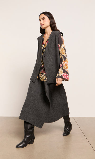 Plümo  Woman in a floral blouse, long gray vest, Eardley Skirt, and black boots stands against a plain wall.