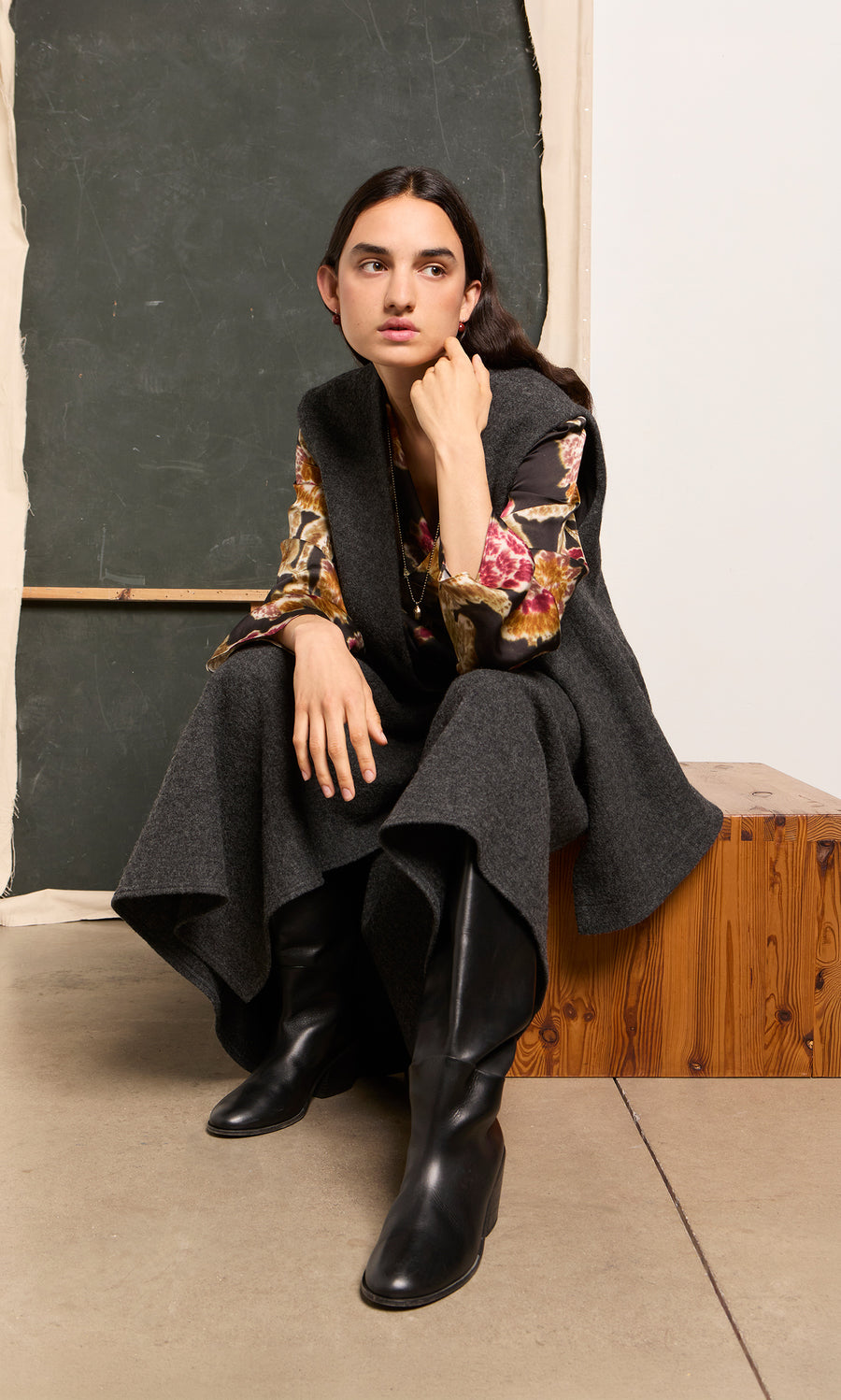 Plümo  Woman in dark gray cape and Tokley Blouse sits on a wooden bench, posing in front of a chalkboard.