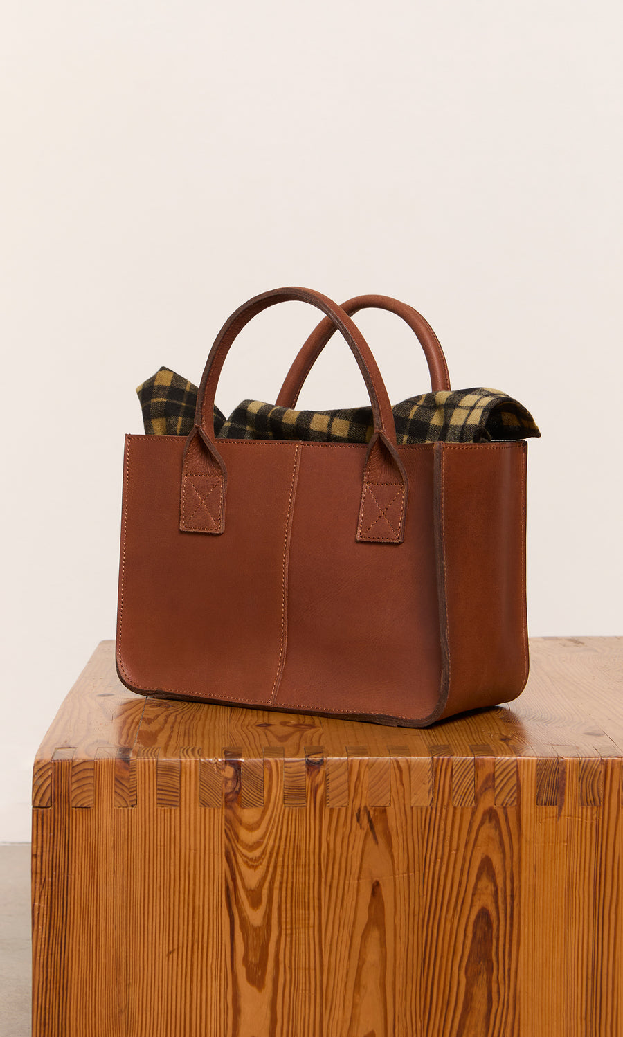 Plümo  Toni Bag: Brown leather tote with plaid lining peeking out, reflecting classic British style on a wooden table.