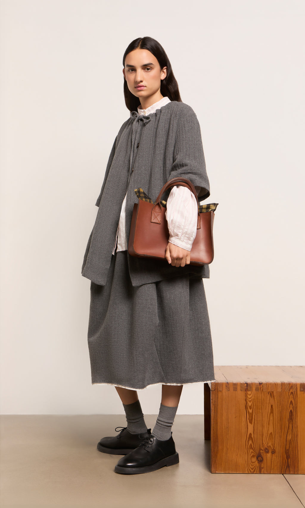 Plümo  Woman with long dark hair holds the Toni Bag, wearing gray and standing by a wooden bench.