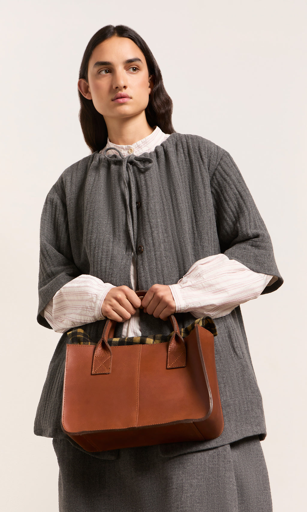 Plümo  Woman in gray holds the Toni Bag, a British design leather tote, looking sideways against a plain background.
