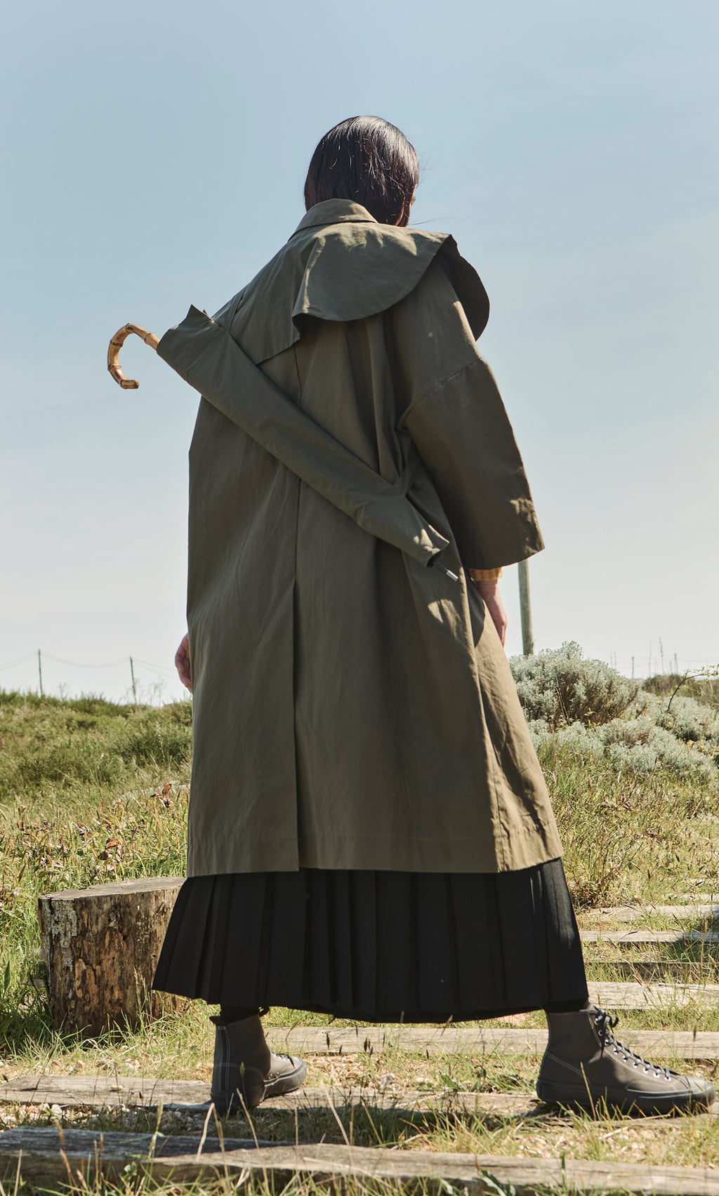 Plümo- A person in a Doherty coat and boots stands outdoors holding a Wax Umbrella Bag on wooden planks.