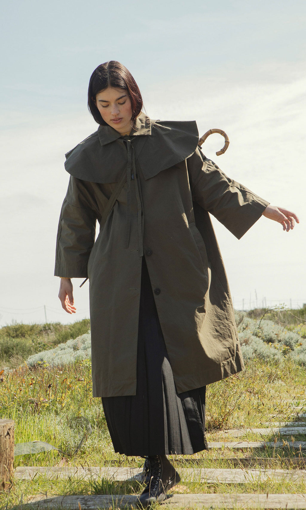 Plümo- Woman in an oversized olive green Doherty coat walks on a stone path, carrying a Wax Umbrella Bag, greenery behind.
