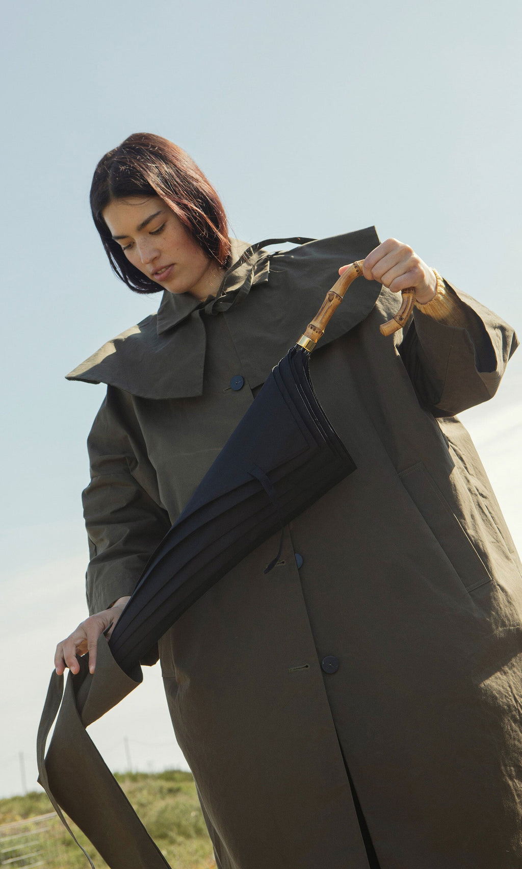 Plümo- A person outdoors under a clear sky holds a closed black umbrella and a Wax Umbrella Bag.